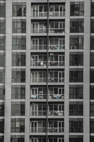 Tall apartment building with multiple floors and balconies filled with household items. Each floor has glass railings and sliding doors, with some balconies displaying clothes hanging to dry.