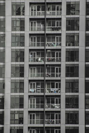 Tall apartment building with multiple floors and balconies filled with household items. Each floor has glass railings and sliding doors, with some balconies displaying clothes hanging to dry.