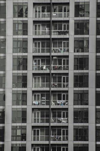 Tall apartment building with multiple floors and balconies filled with household items. Each floor has glass railings and sliding doors, with some balconies displaying clothes hanging to dry.