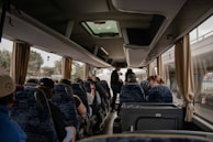 A group of people is seated inside a bus, with several individuals looking out the window and a few standing. The interior features blue patterned seats, beige curtains, and overhead luggage compartments. The bus is parked, as it appears stationary with visible trees and signs outside.