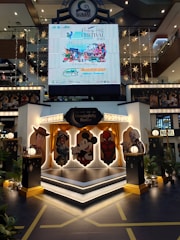A shopping mall interior decorated with a stage featuring cartoon character cutouts, including a cowboy, a female character in traditional attire, a mouse, and a superhero. Above the stage, there's a large digital display promoting the Penang Hill Festival from 21-23 July 2023. The scene is adorned with star-shaped hanging lights and plants, creating a festive atmosphere.