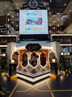 A shopping mall interior decorated with a stage featuring cartoon character cutouts, including a cowboy, a female character in traditional attire, a mouse, and a superhero. Above the stage, there's a large digital display promoting the Penang Hill Festival from 21-23 July 2023. The scene is adorned with star-shaped hanging lights and plants, creating a festive atmosphere.