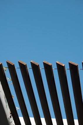 Wooden beams are arranged in a linear pattern against a clear blue sky, creating a striking architectural design. The beams are part of a structure with some greenery visible in the bottom left corner.