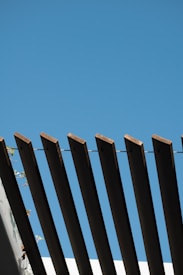 Wooden beams are arranged in a linear pattern against a clear blue sky, creating a striking architectural design. The beams are part of a structure with some greenery visible in the bottom left corner.