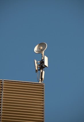 A satellite dish and antenna mounted on the corner of a modern building with slatted panels, set against a clear blue sky.