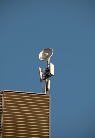 A satellite dish and antenna mounted on the corner of a modern building with slatted panels, set against a clear blue sky.