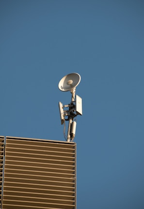 A satellite dish and antenna mounted on the corner of a modern building with slatted panels, set against a clear blue sky.