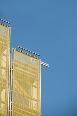 A section of a modern building facade with two vertical yellow mesh panels against a clear blue sky. A small CCTV camera is mounted on one of the panels.