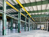 A spacious, industrial warehouse with green steel beams and bright yellow overhead cranes. The floor is concrete and mostly empty, with some machinery and construction material scattered throughout. Large windows allow natural light to fill the interior. Red fire extinguishers are seen along the support columns.