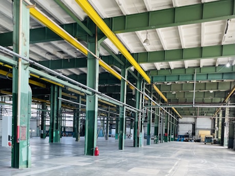 A spacious, industrial warehouse with green steel beams and bright yellow overhead cranes. The floor is concrete and mostly empty, with some machinery and construction material scattered throughout. Large windows allow natural light to fill the interior. Red fire extinguishers are seen along the support columns.