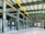 A spacious, industrial warehouse with green steel beams and bright yellow overhead cranes. The floor is concrete and mostly empty, with some machinery and construction material scattered throughout. Large windows allow natural light to fill the interior. Red fire extinguishers are seen along the support columns.