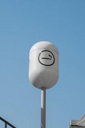A tall, white cylindrical pole with a rounded top displays a smoking symbol against a clear blue sky. The sign is meant to indicate a designated smoking area and features a cigarette icon inside a circle.