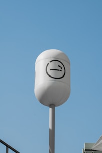 A tall, white cylindrical pole with a rounded top displays a smoking symbol against a clear blue sky. The sign is meant to indicate a designated smoking area and features a cigarette icon inside a circle.