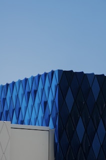 A modern architectural structure featuring a facade with a geometric pattern of blue diamond-shaped panels. The building has a minimalistic and sleek design, with strong lines and a uniform color scheme. The clear blue sky serves as the backdrop.