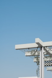 A modern architectural structure featuring a pergola with a minimalist design. The structure includes white beams and metal support, with green plants climbing up and around the frame. The background is an expansive, clear blue sky, creating a contrast with the white and green elements of the structure.