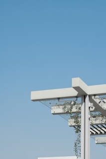 A modern architectural structure featuring a pergola with a minimalist design. The structure includes white beams and metal support, with green plants climbing up and around the frame. The background is an expansive, clear blue sky, creating a contrast with the white and green elements of the structure.