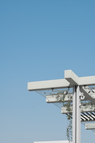 A modern architectural structure featuring a pergola with a minimalist design. The structure includes white beams and metal support, with green plants climbing up and around the frame. The background is an expansive, clear blue sky, creating a contrast with the white and green elements of the structure.
