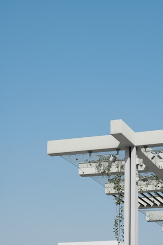 A modern architectural structure featuring a pergola with a minimalist design. The structure includes white beams and metal support, with green plants climbing up and around the frame. The background is an expansive, clear blue sky, creating a contrast with the white and green elements of the structure.