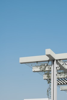 A modern architectural structure featuring a pergola with a minimalist design. The structure includes white beams and metal support, with green plants climbing up and around the frame. The background is an expansive, clear blue sky, creating a contrast with the white and green elements of the structure.