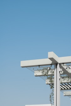 A modern architectural structure featuring a pergola with a minimalist design. The structure includes white beams and metal support, with green plants climbing up and around the frame. The background is an expansive, clear blue sky, creating a contrast with the white and green elements of the structure.