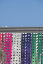 A modern building facade features a series of vertical panels with a metallic, perforated design. The panels display a gradient of colors including pink, white, blue, and green. The sky above is clear and bright blue.