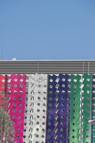 A modern building facade features a series of vertical panels with a metallic, perforated design. The panels display a gradient of colors including pink, white, blue, and green. The sky above is clear and bright blue.
