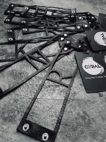 A collection of black, textured, geometric frames are arranged on a stone-like surface. The frames feature metal rivets and are accompanied by black paper tags with branding logos and text.