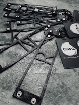A collection of black, textured, geometric frames are arranged on a stone-like surface. The frames feature metal rivets and are accompanied by black paper tags with branding logos and text.