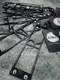 A collection of black, textured, geometric frames are arranged on a stone-like surface. The frames feature metal rivets and are accompanied by black paper tags with branding logos and text.