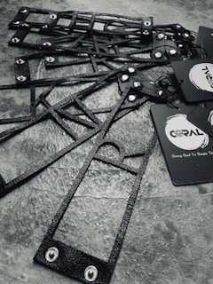 A collection of black, textured, geometric frames are arranged on a stone-like surface. The frames feature metal rivets and are accompanied by black paper tags with branding logos and text.