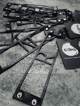 A collection of black, textured, geometric frames are arranged on a stone-like surface. The frames feature metal rivets and are accompanied by black paper tags with branding logos and text.