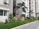 A row of contemporary apartment buildings with tall palm trees and neatly landscaped gardens. The walls are painted in neutral colors and air conditioning units are visible on the exterior. A pedestrian pathway runs alongside small lawns with a variety of green plants and shrubs.