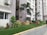A row of contemporary apartment buildings with tall palm trees and neatly landscaped gardens. The walls are painted in neutral colors and air conditioning units are visible on the exterior. A pedestrian pathway runs alongside small lawns with a variety of green plants and shrubs.