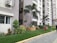 A row of contemporary apartment buildings with tall palm trees and neatly landscaped gardens. The walls are painted in neutral colors and air conditioning units are visible on the exterior. A pedestrian pathway runs alongside small lawns with a variety of green plants and shrubs.
