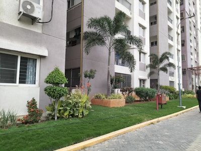 A row of contemporary apartment buildings with tall palm trees and neatly landscaped gardens. The walls are painted in neutral colors and air conditioning units are visible on the exterior. A pedestrian pathway runs alongside small lawns with a variety of green plants and shrubs.