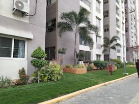 A row of contemporary apartment buildings with tall palm trees and neatly landscaped gardens. The walls are painted in neutral colors and air conditioning units are visible on the exterior. A pedestrian pathway runs alongside small lawns with a variety of green plants and shrubs.