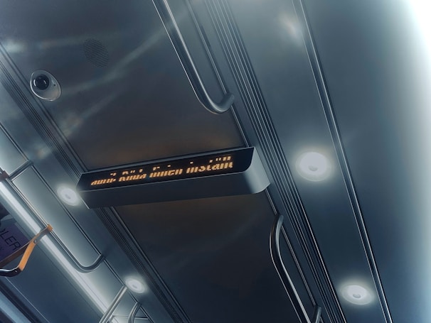 A view of a subway or train interior ceiling showing metal handrails, two spotlights, and a digital display with orange text. A security camera is visible on the ceiling.