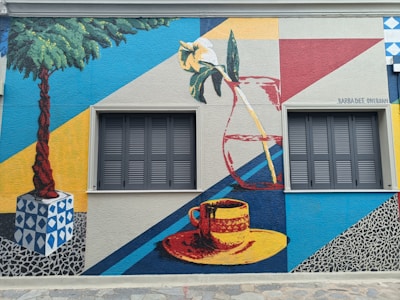 A vibrant mural featuring geometric shapes and vivid colors, depicting a potted palm tree, a flower in a vase, and a cup with a saucer. The artwork incorporates elements like leaves and modern abstract designs, framed by two closed windows with shutters.