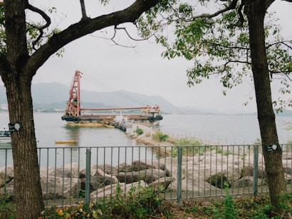 A large barge with a crane is stationed on a calm body of water, with a mountain range in the background. The scene is framed by two trees, and there is a metal fence and rocky shoreline in the foreground. A small yellow boat is floating near the barge.