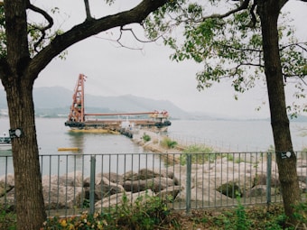 A large barge with a crane is stationed on a calm body of water, with a mountain range in the background. The scene is framed by two trees, and there is a metal fence and rocky shoreline in the foreground. A small yellow boat is floating near the barge.