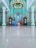 An ornately designed interior of a mosque features a large chandelier with golden accents. The walls are adorned with intricate teal and white patterns, and there are two large grandfather clocks on either side of a beautifully carved wooden pulpit. Five people are seated on the floor, with their reflections visible on the glossy surface.