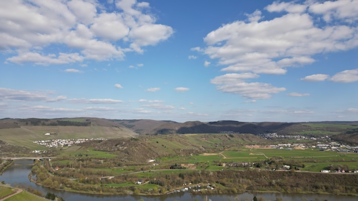 A panoramic view of a landscape featuring rolling hills, a river winding through the foreground, and a small town nestled among the green fields and wooded areas. The sky is partly cloudy with a bright blue background, creating a serene and peaceful scene.