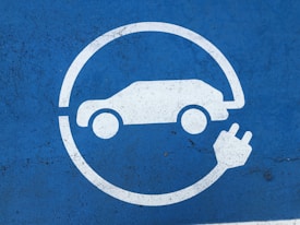 A large white icon depicting a stylized car with a charging plug is centered on a textured blue surface. The image suggests an electric vehicle charging point.