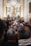 A crowded gathering inside an ornate church with a large altar decorated with flowers. At the center, a statue of a religious figure is elevated, surrounded by columns and elaborate carvings. Many people are present, some taking photos, creating a sense of community and tradition.