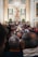 A crowded gathering inside an ornate church with a large altar decorated with flowers. At the center, a statue of a religious figure is elevated, surrounded by columns and elaborate carvings. Many people are present, some taking photos, creating a sense of community and tradition.