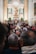 A crowded gathering inside an ornate church with a large altar decorated with flowers. At the center, a statue of a religious figure is elevated, surrounded by columns and elaborate carvings. Many people are present, some taking photos, creating a sense of community and tradition.