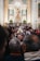 A crowded gathering inside an ornate church with a large altar decorated with flowers. At the center, a statue of a religious figure is elevated, surrounded by columns and elaborate carvings. Many people are present, some taking photos, creating a sense of community and tradition.
