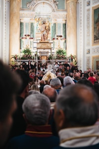 A crowded gathering inside an ornate church with a large altar decorated with flowers. At the center, a statue of a religious figure is elevated, surrounded by columns and elaborate carvings. Many people are present, some taking photos, creating a sense of community and tradition.