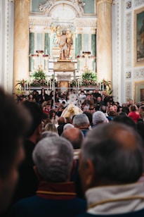 A crowded gathering inside an ornate church with a large altar decorated with flowers. At the center, a statue of a religious figure is elevated, surrounded by columns and elaborate carvings. Many people are present, some taking photos, creating a sense of community and tradition.