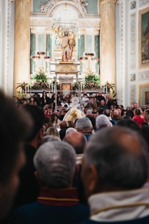 A crowded gathering inside an ornate church with a large altar decorated with flowers. At the center, a statue of a religious figure is elevated, surrounded by columns and elaborate carvings. Many people are present, some taking photos, creating a sense of community and tradition.